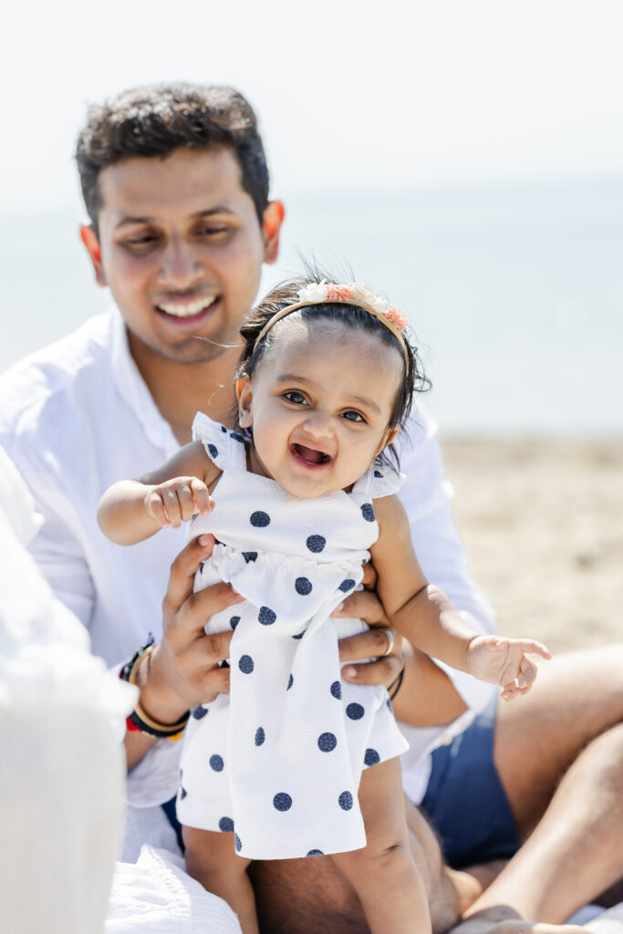 Father sitting down on a beach holding a baby in a standing position as the baby smiles towards the camera during a family photo session. Photo by Tambella Photography