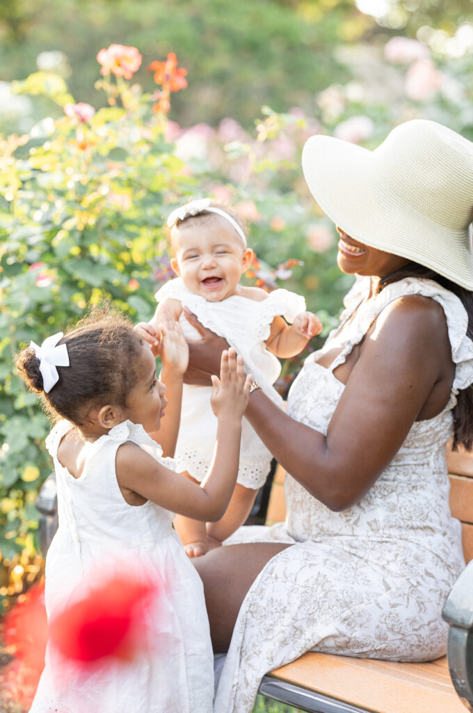 Woman wearing a straw hat sits on a bench in a garden and is surrounded by a baby girl on her lap and a toddler girl standing beside here. They are all smiling and laughing together during a family photography session, photo taken by Tambella Photography