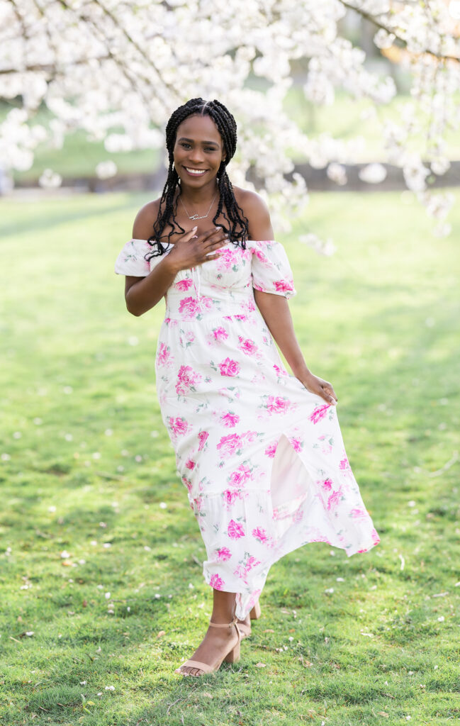 A woman in a white a floral dress standing under a cherry blossom tree.